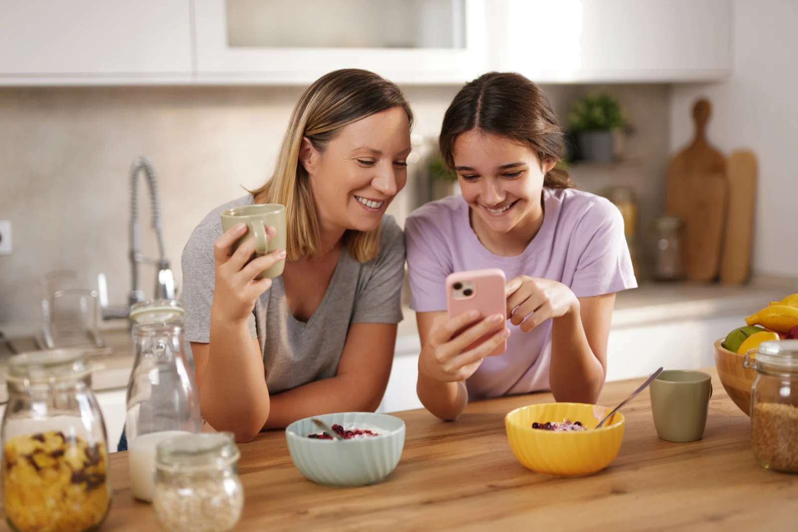 A mother and teenage daughter smiling at a phone together while they eat breakfast